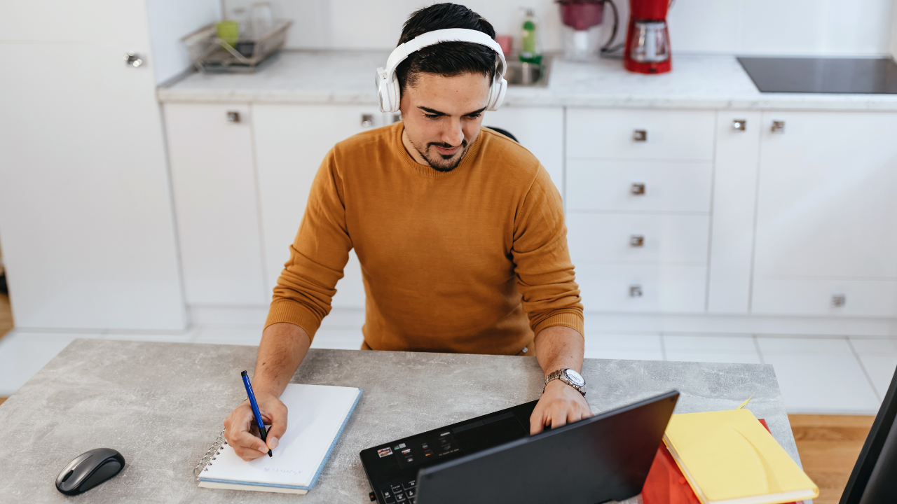 man with headphones using laptop and writing on his notebook